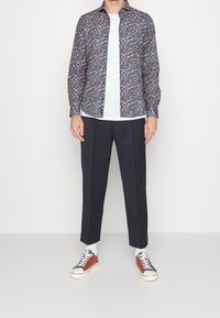 Man in een marineblauwe bloemenoverhemd met lange mouwen over een witte t-shirt, zwarte broek, witte sokken en multicolor casual sneakers staande tegen een witte achtergrond.