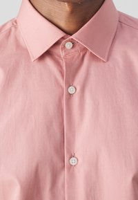 Lichtroze overhemd met knoopsluiting, voorzien van een spread collar en witte kunststof knopen. De stof ziet er glad uit met een licht gekreukt textuur.