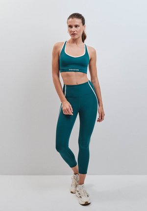 Donna in piedi con reggiseno sportivo turchese e leggings con dettagli a strisce bianche e sneakers beige su sfondo grigio chiaro uniforme.