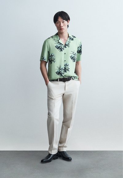 Νεαρός άνδρας όρθιος με τα χέρια στις τσέπες, φορώντας πράσινο φλοράλ κοντομάνικο πουκάμισο, μπεζ παντελόνι, μαύρη ζώνη και μαύρα παπούτσια σε γκρι πάτωμα.