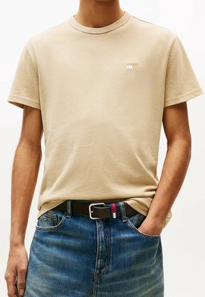 Man draagt beige T-shirt met korte mouwen en klein logo op de borst, blauwe jeans en zwarte riem met rood-wit-blauw gestreept detail.
