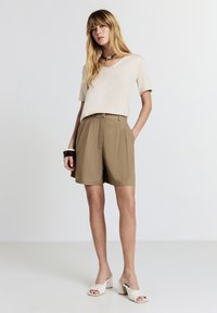Giovane donna con top beige a maniche corte, pantaloni corti khaki a vita alta, sandali bianchi con tacco e bracciale geometrico che si trova davanti a uno sfondo semplice.