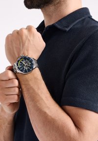 Nerezové chronograf hodinky s tmavě modrým ciferníkem, stříbrnými akcenty, tachymetrickou škálou a texturovaným náramkem, nošené na mužském zápěstí.