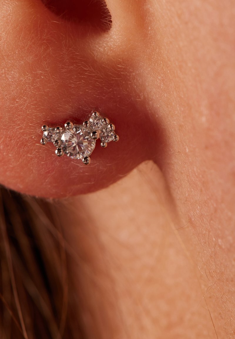 Pendientes de Stud brillantes con gemas claras en un diseño de racimo, engastados en un metal de tono plateado, colocados sobre la piel.