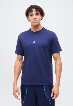 Mladý muž stojí, nosí tmavomodré tričko Jordan s bielym logom a zodpovedajúce šortky so svetlomodrým lemovaním, jednoduché svetlé pozadie.