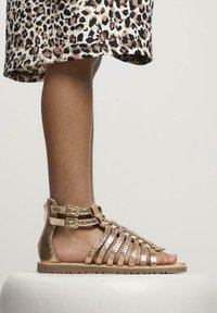 Sandalias metálicas doradas con tiras, acabado texturizado, correas en el tobillo con hebilla y suela plana. Llevadas con una falda de estampado de leopardo hasta la rodilla.