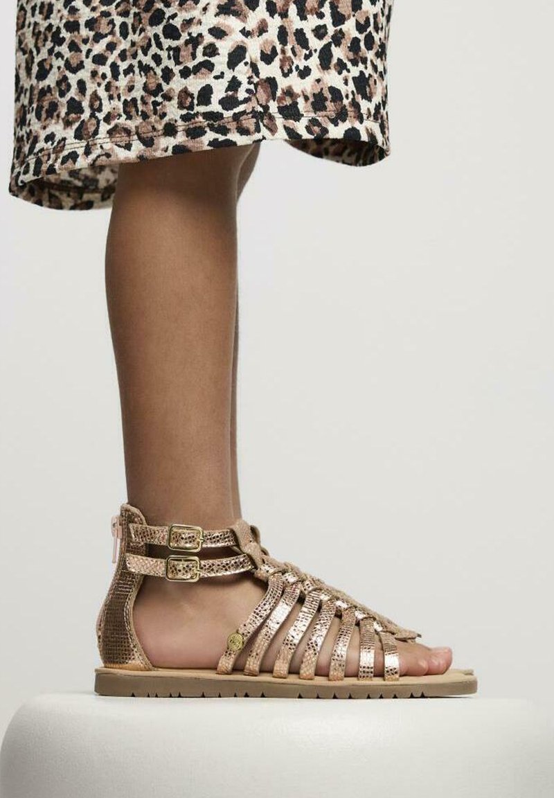Sandalias metálicas doradas con tiras, acabado texturizado, correas en el tobillo con hebilla y suela plana. Llevadas con una falda de estampado de leopardo hasta la rodilla.