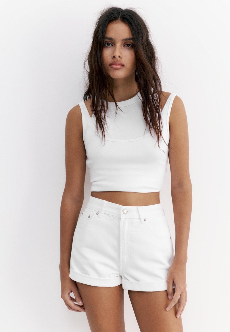 Junge Frau mit langen dunklen Haaren, die ein weißes ärmelloses Crop-Top und weiße hoch taillierte Shorts mit aufgerolltem Saum trägt, steht vor einem schlichten weißen Hintergrund.