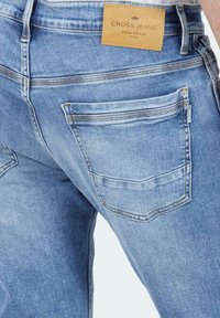 Jasnoniebieskie dżinsy o teksturowanym wykończeniu, z bocznymi i tylnymi kieszeniami oraz brązową metką z napisem "CROSS JEANS" na pasku.