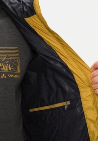 Interno della giacca reversibile nera e oro; presenta una tasca con zip e una superficie liscia. Sotto, una maglietta grigia con una grafica di montagna.