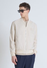Licht beige bomberjack met een ritssluiting aan de voorkant, geribbelde manchetten en een klein logo-detail. Gladde textuur, comfortabele fit, gestyled met donkere broeken en een zonnebril.