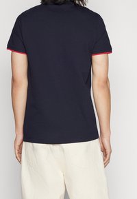 Polo di colore blu navy con rifiniture rosse sulle maniche, maniche corte e vestibilità classica, realizzato in morbido tessuto di cotone, design minimalista.