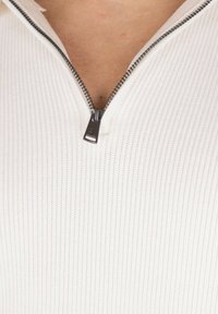 Maglia bianca a maniche lunghe con costine e mezza zip al colletto. Il materiale è tessuto e aderente, con un tirante metallico per la zip.