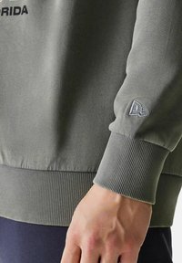 Nahaufnahme einer Hand neben einem gerippten Bündchen eines grauen Sweatshirts mit einem kleinen gestickten Logo und teilweise schwarzem Text auf dem Stoff.