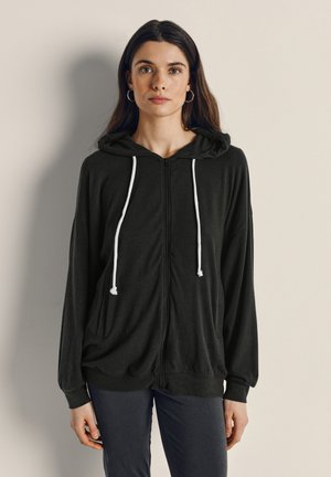 Ung kvinna med långt mörkt hår, klädd i en svart dragkedjehoodie med vita snören, stående mot en ljust färgad vägg.