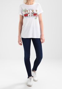 Weißes T-Shirt mit goldener und schwarzer gedruckter Schrift sowie floralen Akzenten; kombiniert mit dunkelblauen engen Jeans und weißen Sportschuhen.