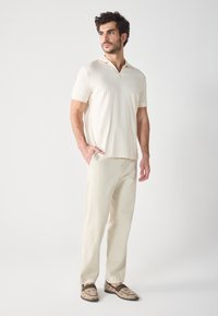 Cremfarbener, strukturierter Poloshirt mit kurzen Ärmeln, kombiniert mit hellbeigen Hosen und gemusterten braunen Schuhen. Schlichte, klassische Passform.