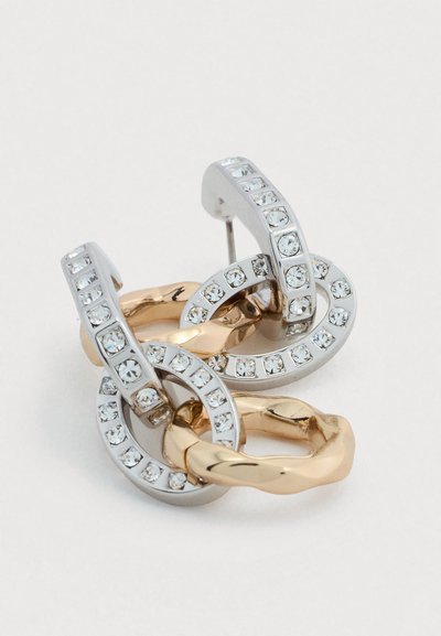 Σκουλαρίκια κρίκοι με διαπλεκόμενο σχεδιασμό από μέταλλο σε ασημένιο και χρυσό χρώμα με κρυστάλλινες λεπτομέρειες. Ομαλές και υφασμένες επιφάνειες δημιουργούν αντίθεση.