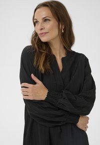 Čierna blúzka s dlhým rukávom a vertikálnou rebrovanou textúrou, so zaobleným výstrihom a zhrnutými manžetami. Hladká tkanina s voľným strihom.