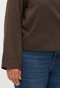 Brown textured top with long sleeves, featuring a subtle woven pattern. Paired with blue denim jeans. Focus on the garment's material.