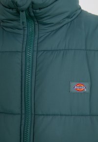 Vadderad jacka i teal med hög krage, framdragkedja och logotyp-patch. Strukturerad tyg med kviltade detaljer och kontrasterande dragkedja.