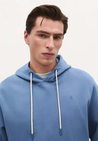 Lichtblauwe hoodie van zacht materiaal met een voorzak, verstelbare trekkoorden en een klein logo op de borst.