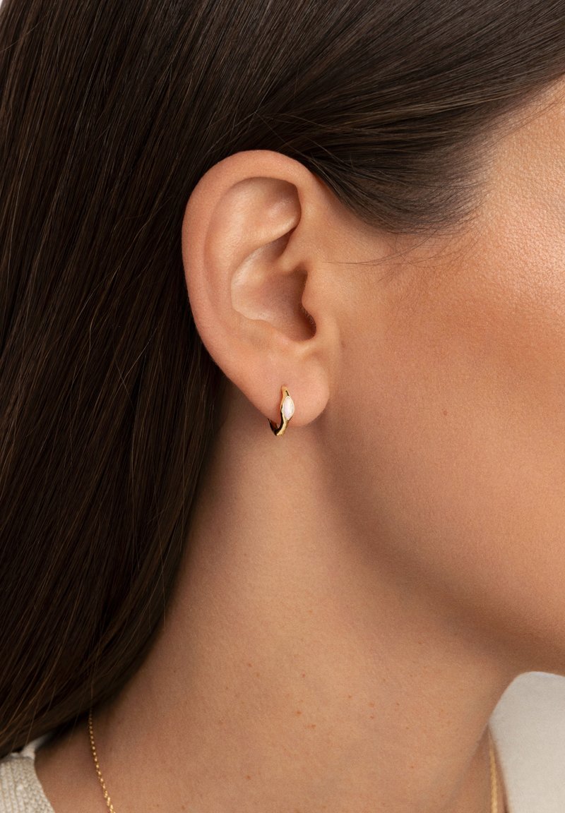 Pendiente de aro dorado con ligera curva y un acento de piedra blanca, exhibido en el lóbulo de la oreja con piel suave y cabello oscuro de fondo.
