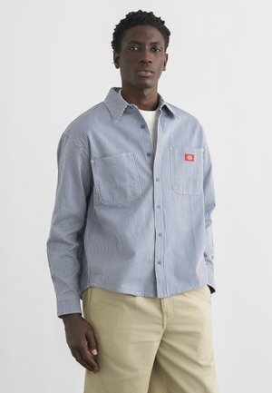 Homme portant une chemise à rayures bleu et blanc boutonnée par-dessus un t-shirt blanc et un pantalon beige, debout devant un fond clair uni.