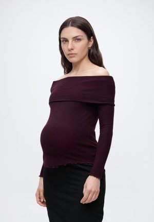 Femme enceinte aux longs cheveux foncés portant un haut bordeaux décolleté épaules et une jupe noire, debout devant un fond blanc uni.