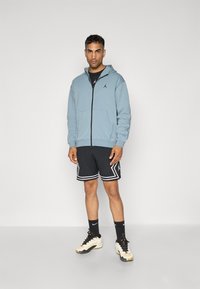 Lyseblå zip-up hoodie med sort logo, parret med sorte mesh shorts med hvide accenter og et lagdelt look. Sorte atletiske sko.
