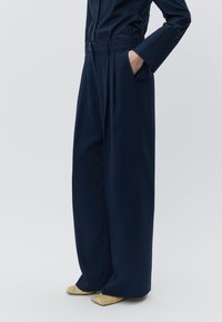 Calças largas azul-marinho com pregas, feitas de tecido suave. Apresentam um cinto na cintura e bolsos laterais, combinadas com sapatos beges pontiagudos.