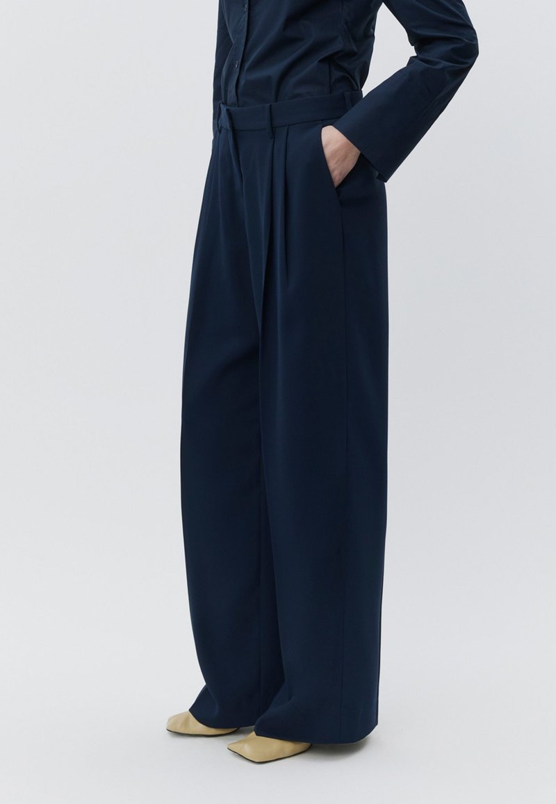 Calças largas azul-marinho com pregas, feitas de tecido suave. Apresentam um cinto na cintura e bolsos laterais, combinadas com sapatos beges pontiagudos.