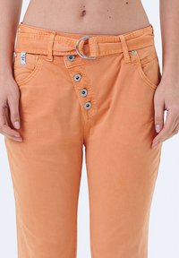 Pantalons en coton orange avec une taille mi-haute, fermeture à bouton en métal et une boucle de ceinture ronde argentée. Possède des poches latérales et des accents cousus.