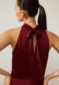 Vinrød halterdragt med knyttet hals, åben ryg, ribbet tekstur og tætsiddende silhuet, der fremhæver krave- og skulderdesignet.