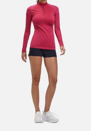 Frau trägt ein enganliegendes langärmeliges pinkes Sportoberteil, schwarze Shorts und weiße Laufschuhe, steht vor einem einfarbigen Hintergrund.