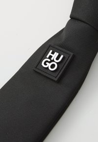 Cratcă neagră din material texturat, cu un eticheta rectangulară pe care este gravat cu litere albe cuvântul "HUGO".
