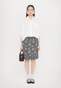 Hvit oversized skjorte med knapper, grå skjørt med blomsterdekorasjoner, brun handveske og brune sko med stropper; ensfarget bakgrunn.