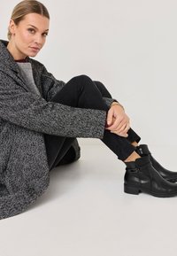 Botas de cuero negro hasta el tobillo con cremallera lateral y correa ajustable. Abrigo gris de espiga con mangas amplias y jeans negros ajustados.