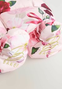 Rosa blomstertrykt stof med broderet gyldent skrift, der står "BAKER." Designet har forskellige blomster og bånd, hvilket skaber et livligt udseende.