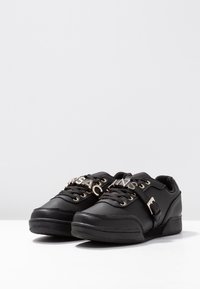 Baskets en cuir noir avec semelle plate, accessoires en ton doré, lacets et une sangle avec une marque décorative à l'avant.