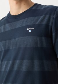 Granatowa koszulka z krótkim rękawem w poziome paski w różnych odcieniach. Posiada okrągły dekolt oraz haftowane logo "Barbour" na piersi.