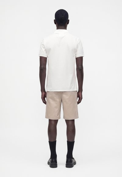 Homme debout, de dos, portant une chemise blanche à manches courtes, un short beige, des chaussettes noires et des chaussures noires sur fond blanc.