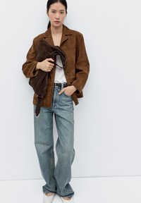 Brązowa zamszowa kurtka na jasnoszarej koszuli, zestawiona z luźnymi niebieskimi dżinsami i białymi butami, trzymająca brązową zamszową torbę. Minimalistyczne tło.