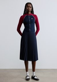 Tumepunast sinisest rippdetailiga lukuga pusa, mille varrukad on marooni värvi, koos volditud tumepunase seelikuga. Välimust täiustavad mustad kingad kuldsete detailidega ja valged sokid.