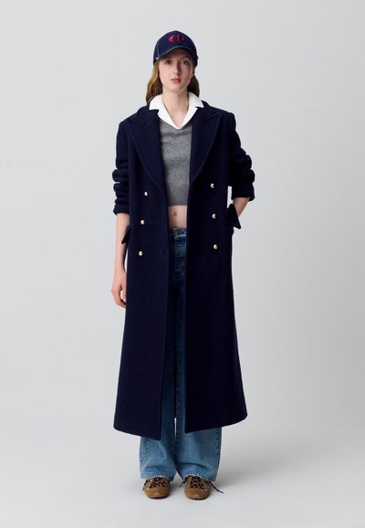 Cappotto di lana blu navy con bottoni dorati doppi, abbinato a un maglione grigio, jeans a palazzo blu e un berretto navy.