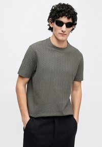 Cămașă gri cu mâneci scurte, cu o suprafață texturată și modelată. Modelul poartă ochelari de soare negri, în picioare cu mâinile în buzunare.