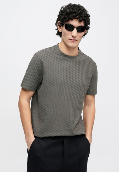 Pilka trumpomis rankovėmis palaidinė su tekstūruota, raštuota paviršiaus danga. Modelis dėvi juodus akinius nuo saulės, stovi su rankomis kišenėse.