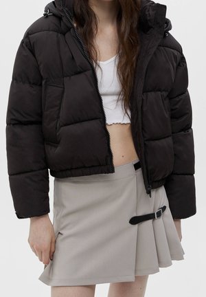 Doudoune noire avec capuche, coupe carrée, texture matelassée et fermeture éclair à l'avant. Assortie à une jupe plissée gris clair, avec un détail de boucle.