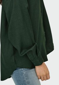 Dark green button-up shirt with a textured fabric, loose fit, and long sleeves, paired with light blue jeans, viewed from the side.