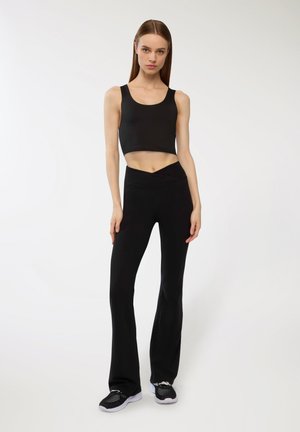 Top corto nero e pantaloni a vita alta svasati neri, entrambi realizzati in tessuto morbido. Modello che indossa scarpe sportive nere con dettagli in rete.